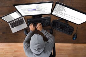 An image featuring a person wearing a hoodie and using multiple computers and a laptop to hack representing a hacker